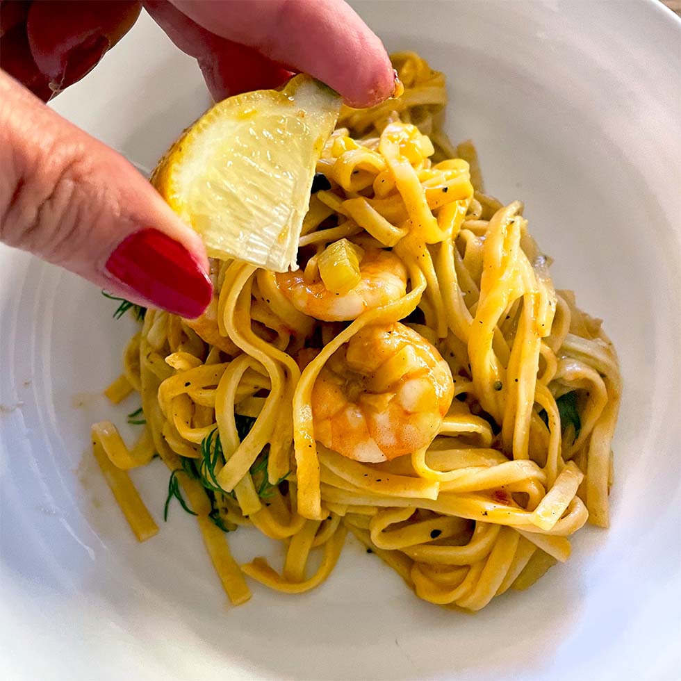 A hand with red nail varnish squeezes a lemon wedge over a plate of prawn pasta with yellow sauce, garnished with fresh herbs.