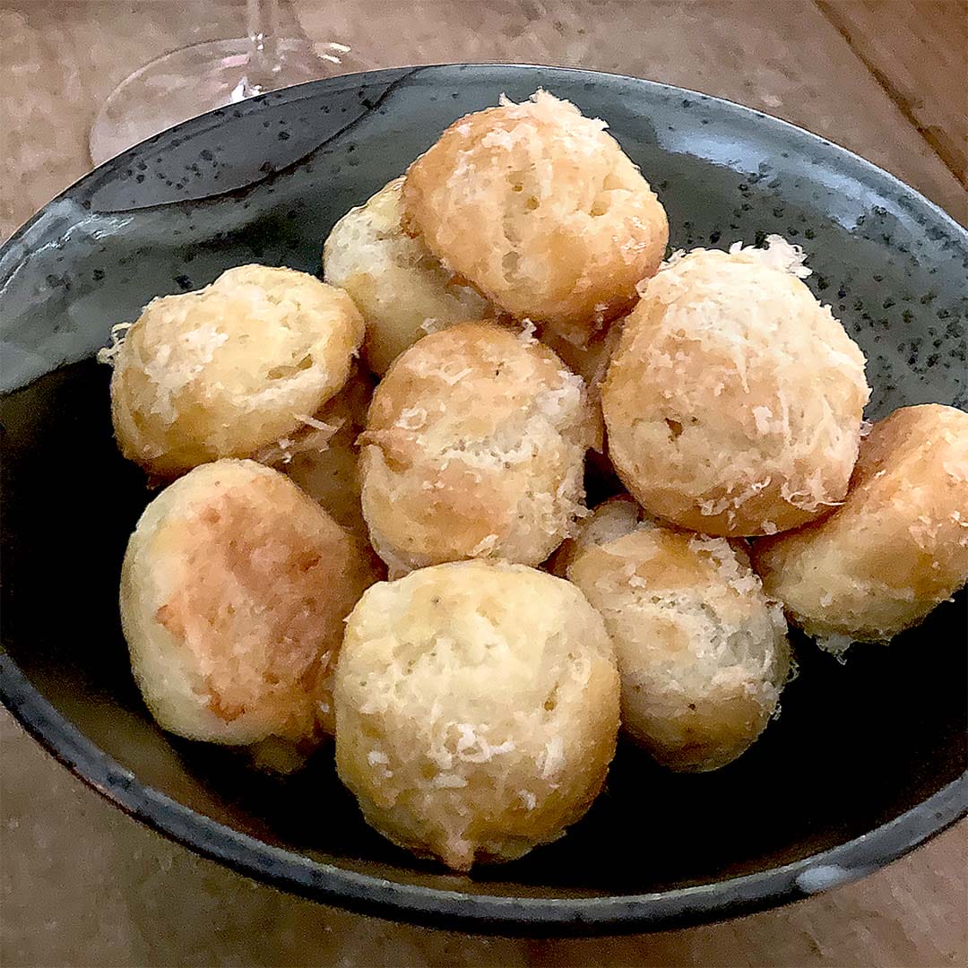 A dark bowl filled with round, golden-brown cheese puffs topped with grated cheese, sitting on a wooden surface.
