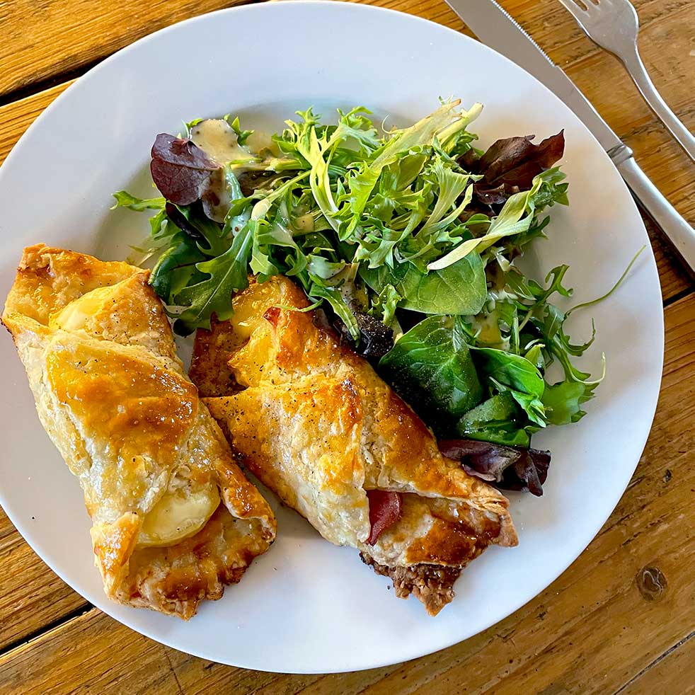 A white plate with two golden, flaky pastry rolls filled with cheese and meat, served alongside a fresh mixed green salad, set on a wooden table with a fork and knife.