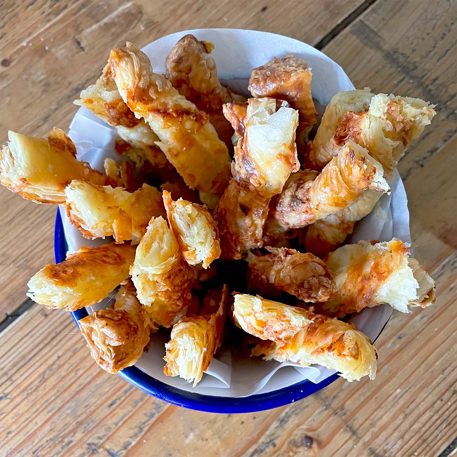 A bowl lined with greaseproof paper filled with golden, flaky cheese straws made from puff pastry, arranged upright on a wooden table.