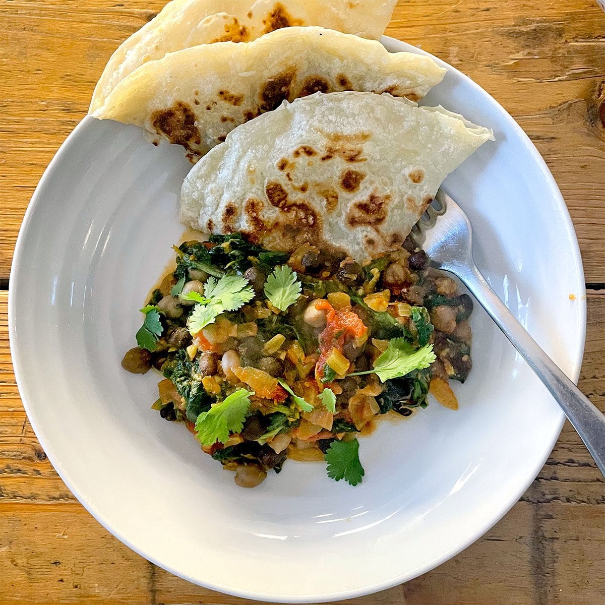 A white plate with chickpea and spinach curry garnished with coriander, served with two pieces of flatbread. A spoon rests on the plate, which is set on a wooden table.