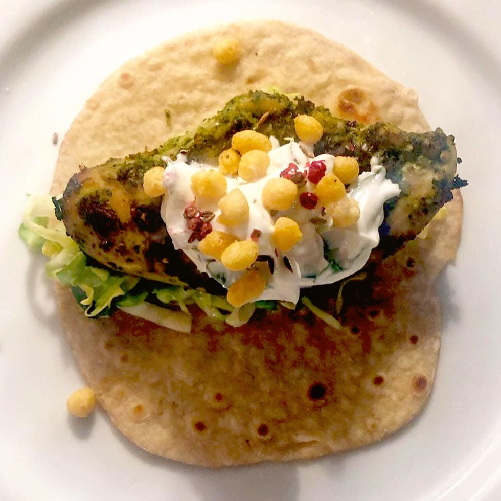 A wholemeal tortilla topped with grilled chicken, shredded lettuce, a dollop of soured cream, crispy round snacks, and red seasoning, served on a white plate.