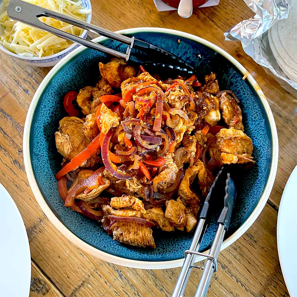 A bowl of cooked chicken strips with red onions and red peppers, served in a blue bowl with tongs. Grated cheese and tortillas are visible nearby on a wooden table.