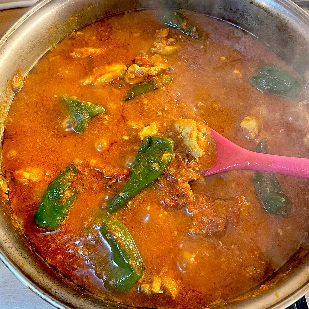 A pot of red, spicy stew with chunks of meat and whole green peppers, being stirred with a pink spoon. Steam rises from the hot, simmering dish.