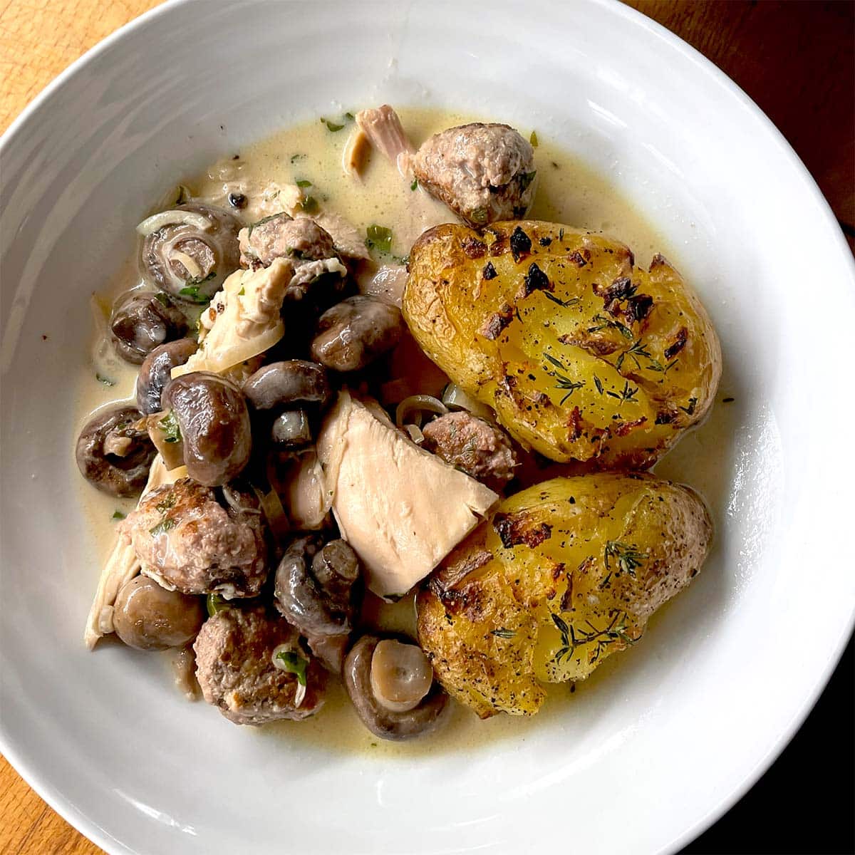 A white bowl contains creamy chicken and mushroom stew with herbs, meatballs, and two crispy, golden roasted smashed potatoes on the side.