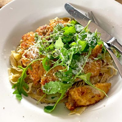A white plate with pasta topped with crispy breaded chicken, fresh rocket, and grated cheese, with a fork and knife resting on the plate.