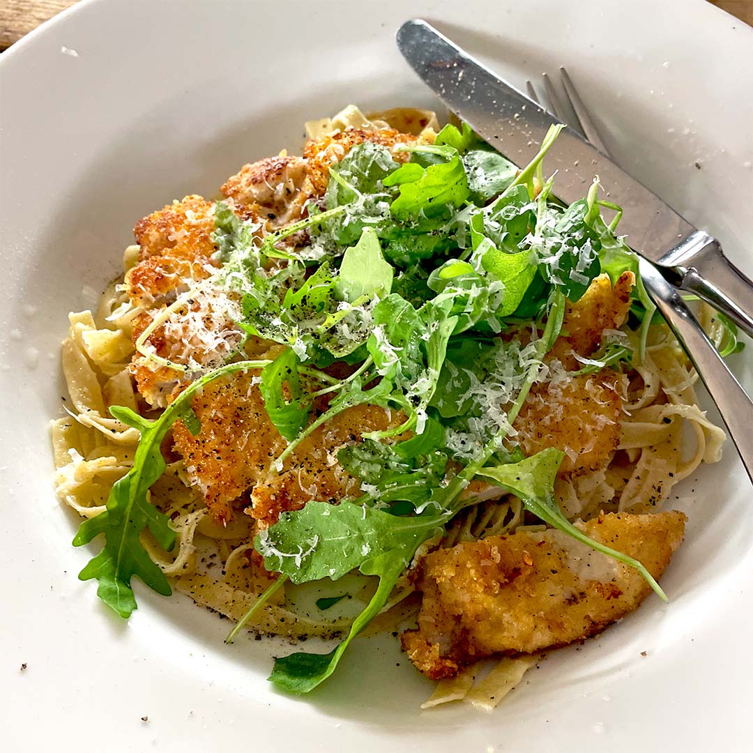 A white plate with pasta topped with crispy breaded chicken, fresh rocket, and grated cheese, with a fork and knife resting on the plate.