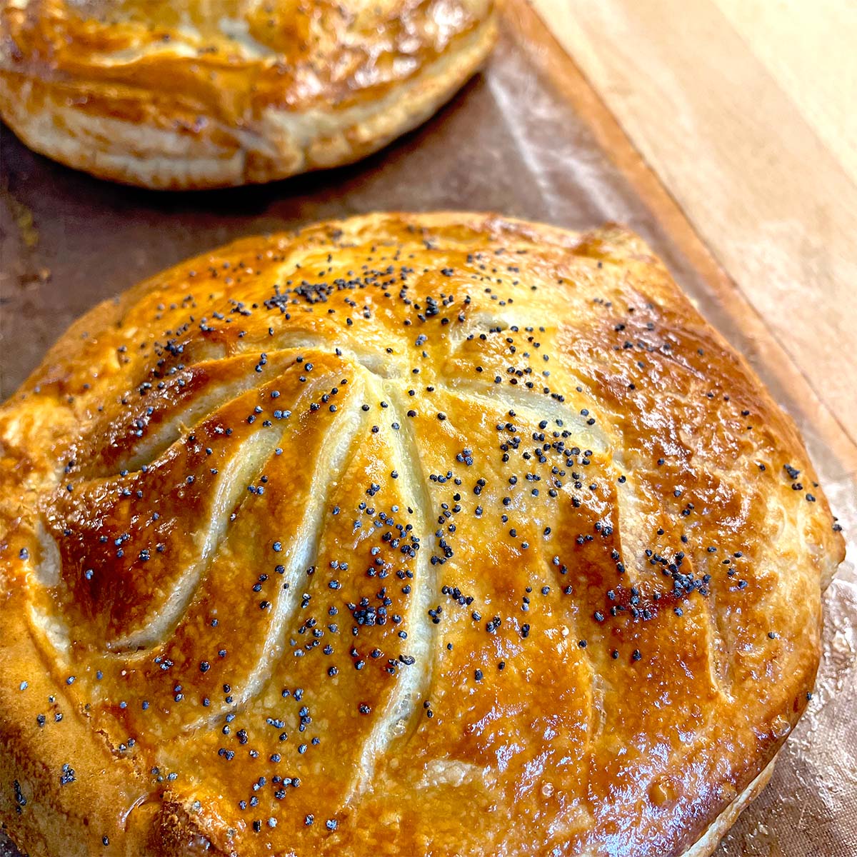 Golden-brown, round pastry topped with poppy seeds and decorative scored lines, resting on a baking tray. Another similar pastry is visible in the background.