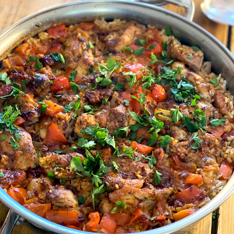 A frying pan filled with arroz con pollo, showing seasoned chicken, rice, tomatoes, red peppers, and fresh parsley on top. The dish looks freshly cooked and colourful.