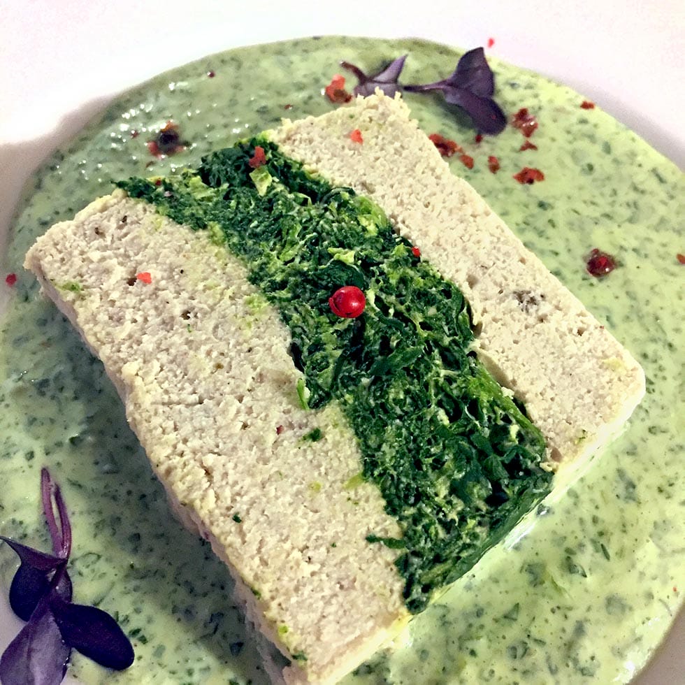 A rectangular slice of pâté with a green spinach centre is served on a creamy green herb sauce, garnished with pink peppercorns and small purple microgreens.