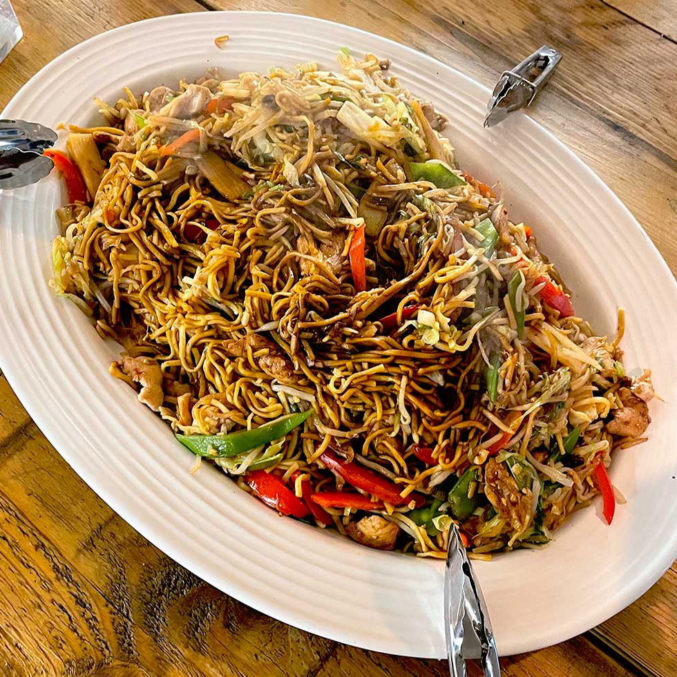 A large oval platter of stir-fried noodles with vegetables, chicken, and red and green peppers is placed on a wooden table, with serving tongs on each end of the dish. Steam rises from the hot food.