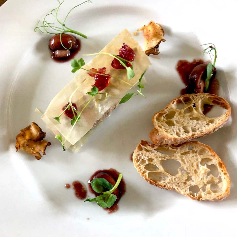 A gourmet dish presented on a white plate with two slices of crusty bread, a rectangular portion of food topped with microgreens and sauce, garnished with mushrooms and olives, and artistic sauce drizzles.