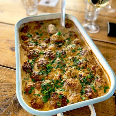 A baking dish filled with creamy meatballs garnished with chopped parsley, placed on a wooden table next to a glass of white wine and some drinking glasses.