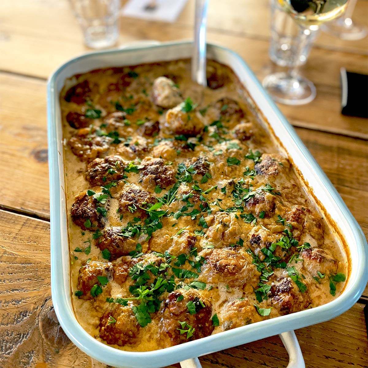 A baking dish filled with creamy meatballs garnished with chopped parsley, placed on a wooden table next to a glass of white wine and some drinking glasses.