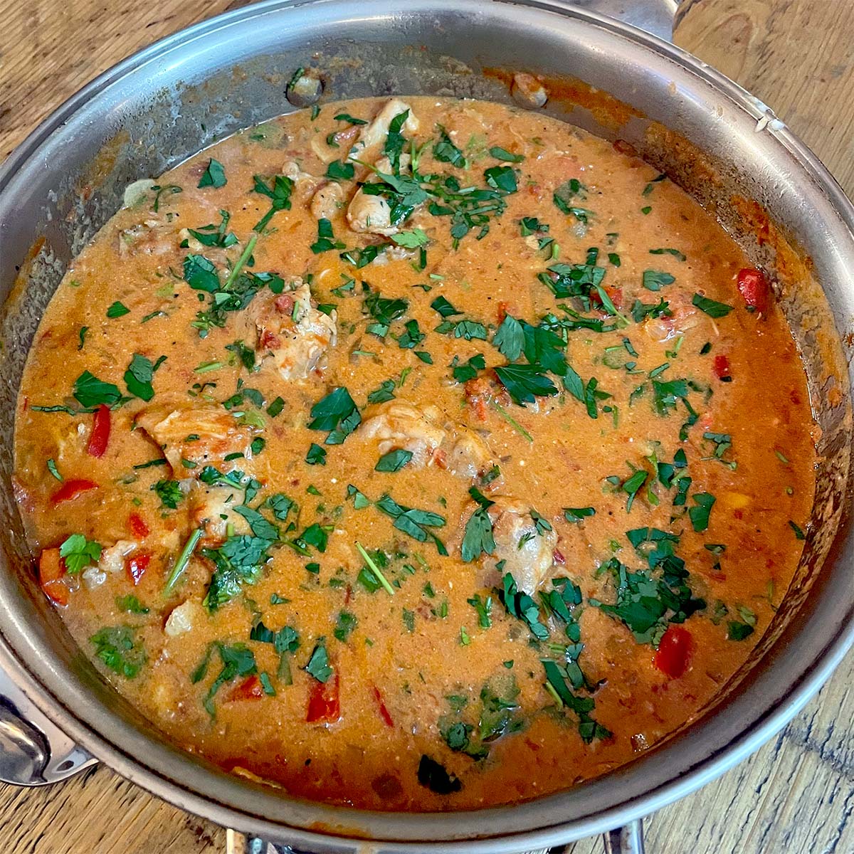 A metal pan filled with creamy, orange-coloured chicken stew, garnished with chopped fresh parsley and pieces of red pepper, sitting on a wooden surface.