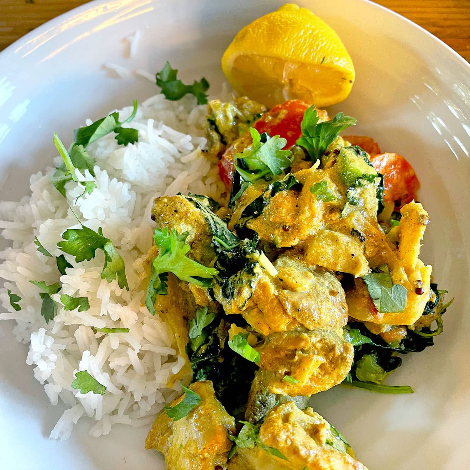 A white plate with steamed white rice, creamy yellow curry with chicken, spinach, tomatoes, and fresh coriander, garnished with a lemon wedge.