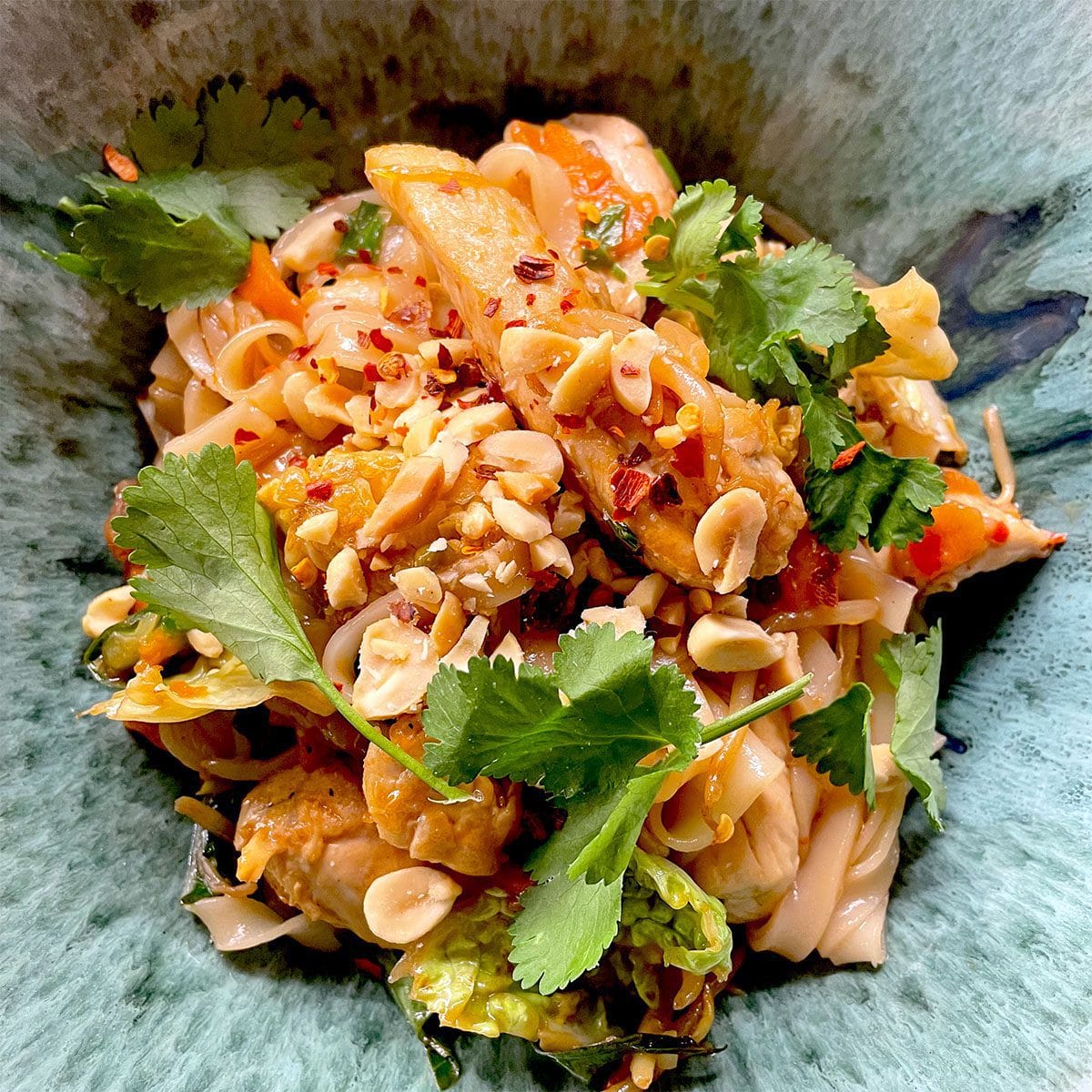 A close-up of a vibrant noodle dish topped with chopped peanuts, fresh coriander, red chilli flakes, and pieces of chicken, served in a green bowl.