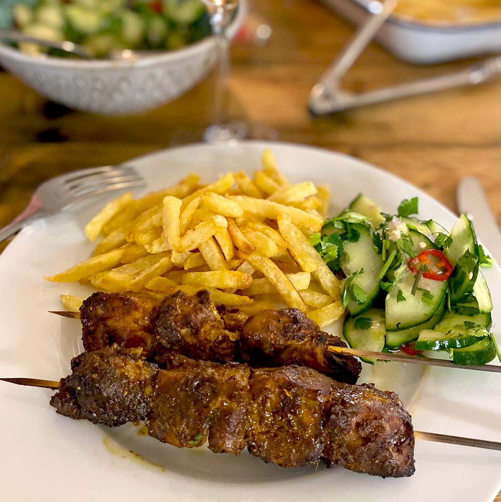 A plate with two grilled meat kebabs, a serving of golden chips, and a fresh cucumber salad garnished with herbs and red chilli slices. A fork and knife rest on the table beside the plate.
