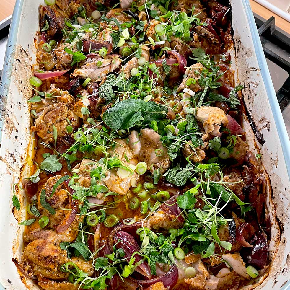 A baked dish filled with roasted chicken pieces, red onions, and herbs like coriander and mint, garnished with sliced spring onions and microgreens. The food is in a white rectangular baking dish on a hob.