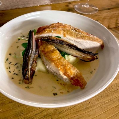A white bowl with roasted chicken breast on creamy mashed potato, topped with grilled vegetables and a light herb sauce, served on a wooden table.