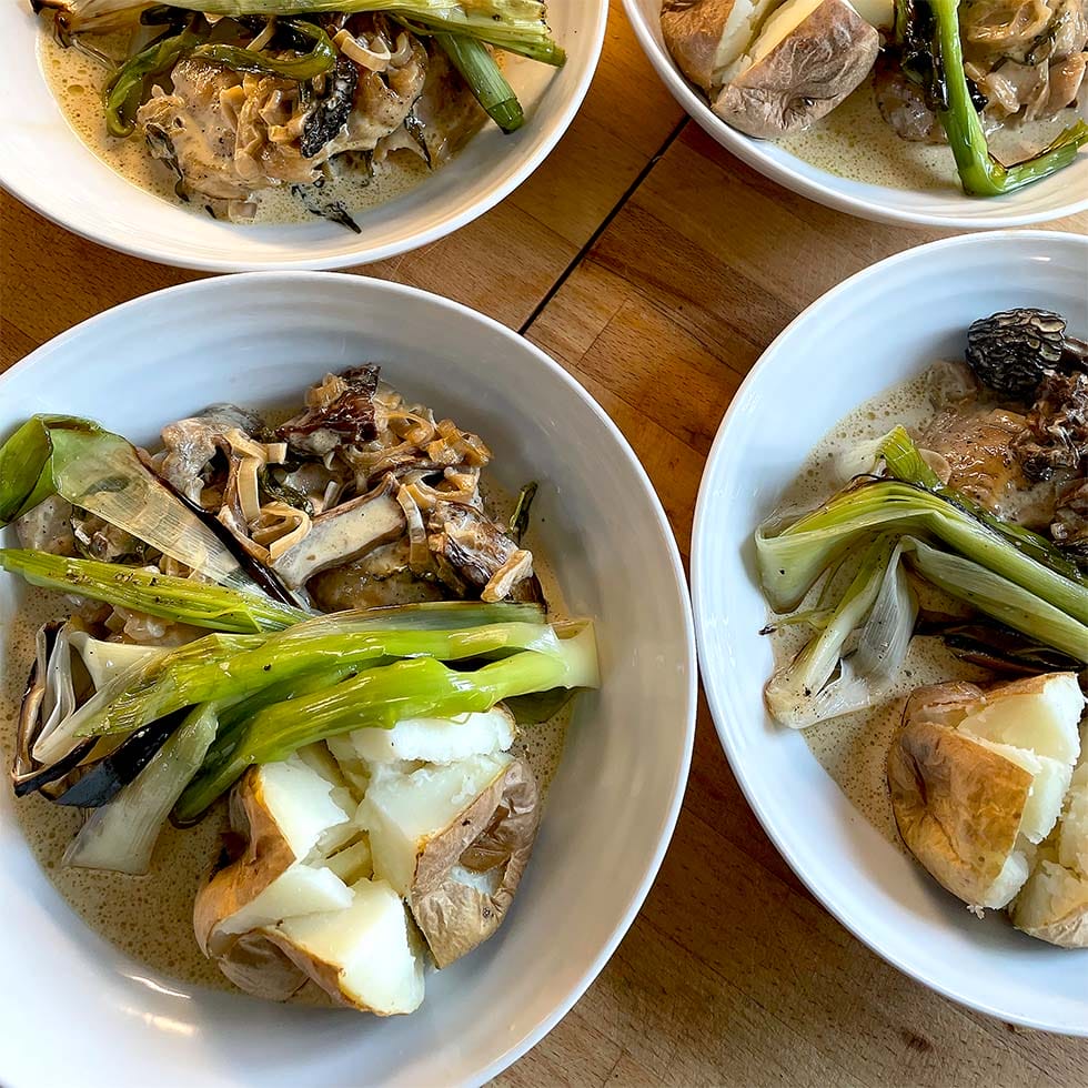 Four white bowls filled with baked potatoes, grilled spring onions, mushrooms, and creamy sauce are arranged on a wooden surface. The dish appears hearty and rustic, featuring roasted and sautéed vegetables.