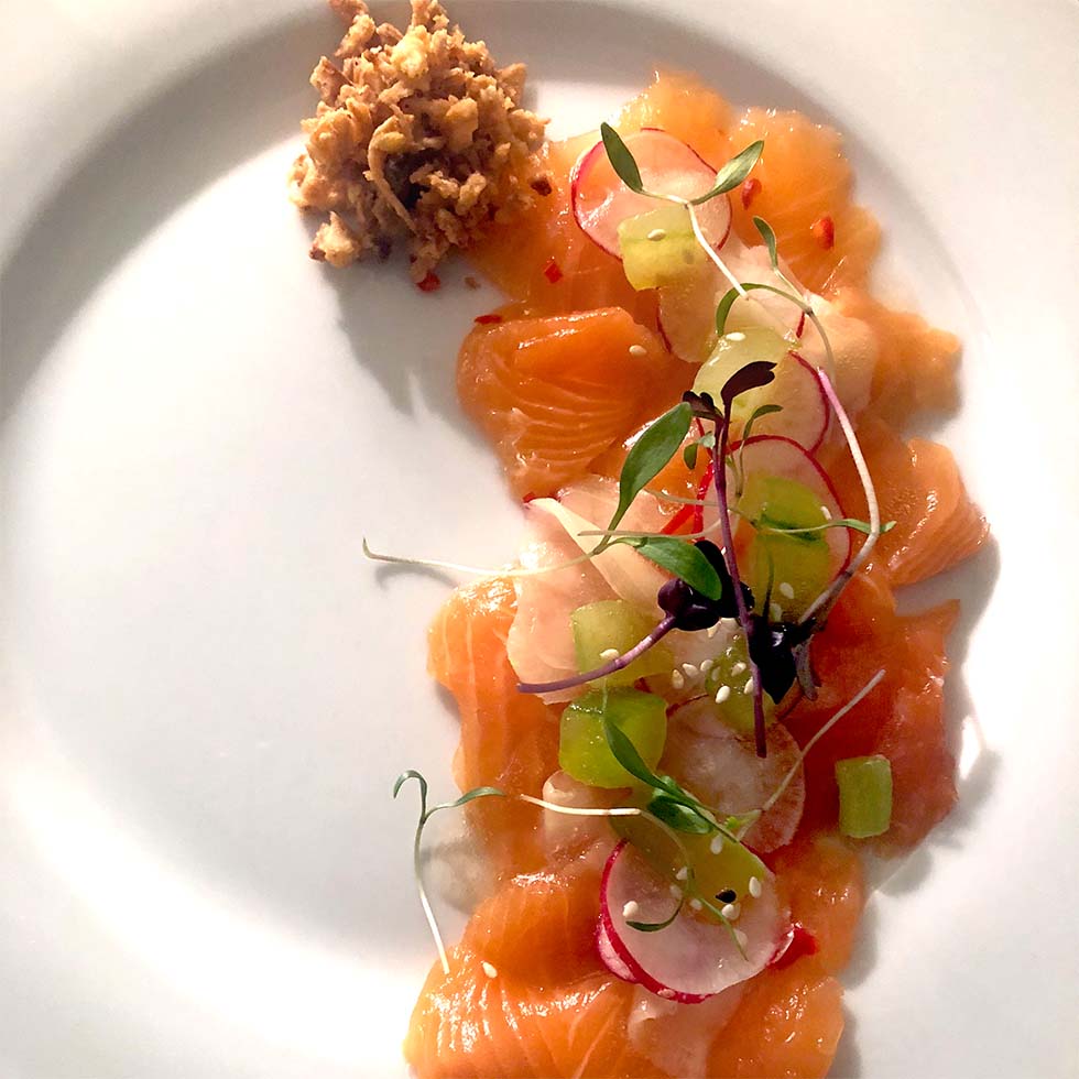 Thin slices of raw salmon arranged on a white plate, garnished with microgreens, radish slices, cucumber, and a small pile of crumbled topping on the side.