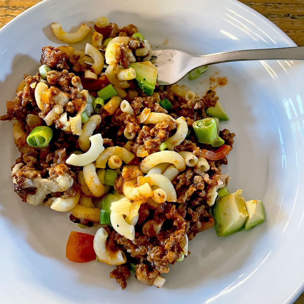 A white plate holds a serving of macaroni pasta mixed with minced beef, diced spring onions, avocado, tomato, and melted cheese. A fork rests on the edge of the plate.