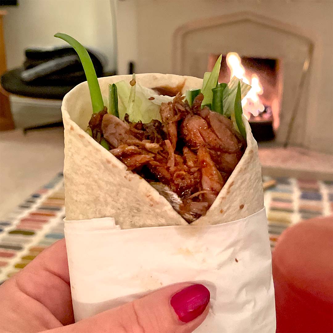 A hand with red nail varnish holds a wrap filled with shredded meat, lettuce, and cucumber. A fireplace burns in the background, creating a cosy indoor atmosphere.