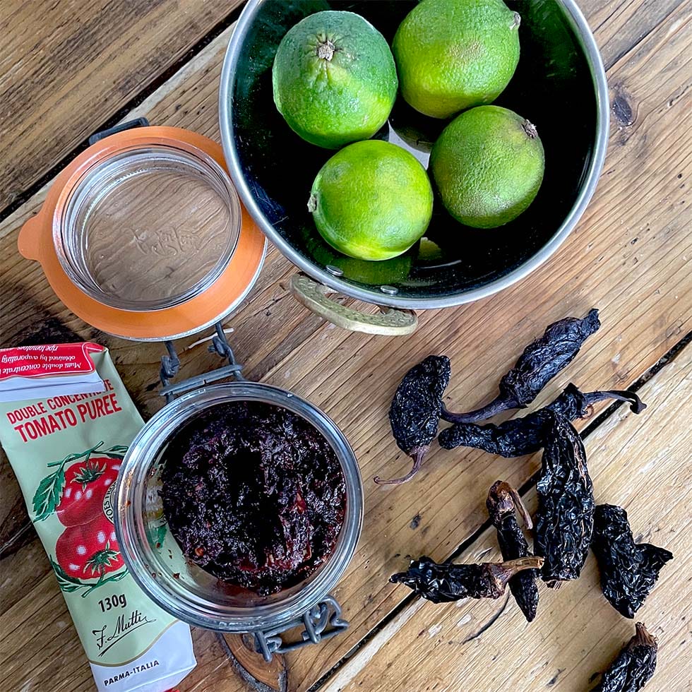 A bowl of three limes, a glass jar of dark paste, dried chillies, a tube of tomato purée, and an empty jar are arranged on a wooden surface.