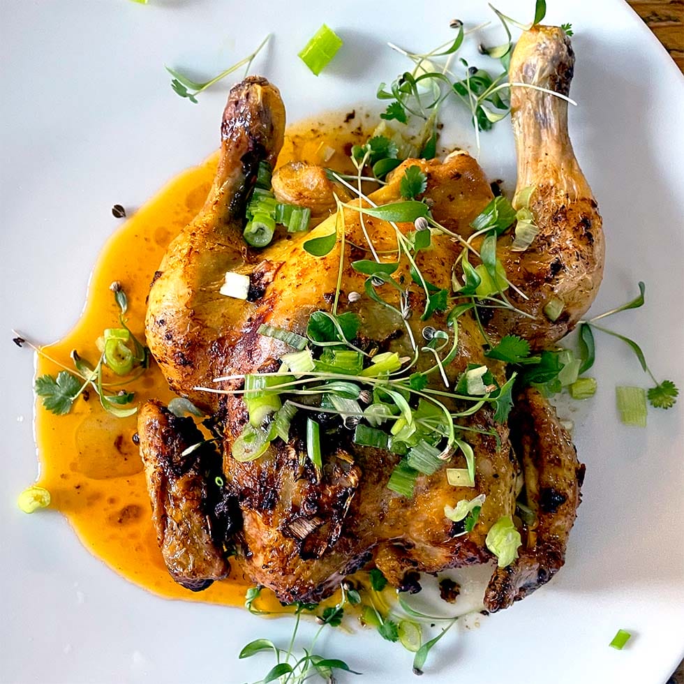 Grilled or roasted half chicken served on a white plate, topped with fresh microgreens, chopped spring onions, and herbs, with a pool of orange-brown sauce underneath.