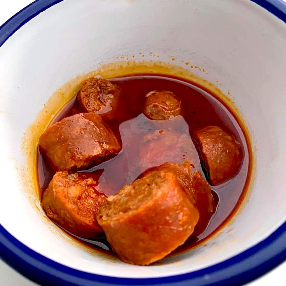 A white bowl with a blue rim contains several pieces of sausage in a rich, oily red sauce, likely a spicy or tomato-based stew.
