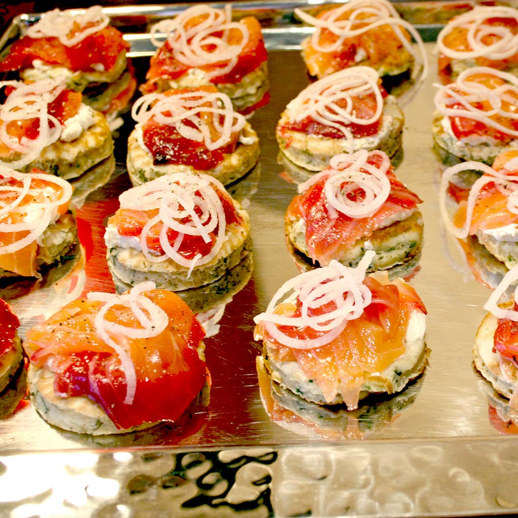 Rows of small, round appetisers on a metal tray, topped with cream, slices of smoked salmon, and thinly sliced onions. The appetisers are neatly arranged and garnished for serving.