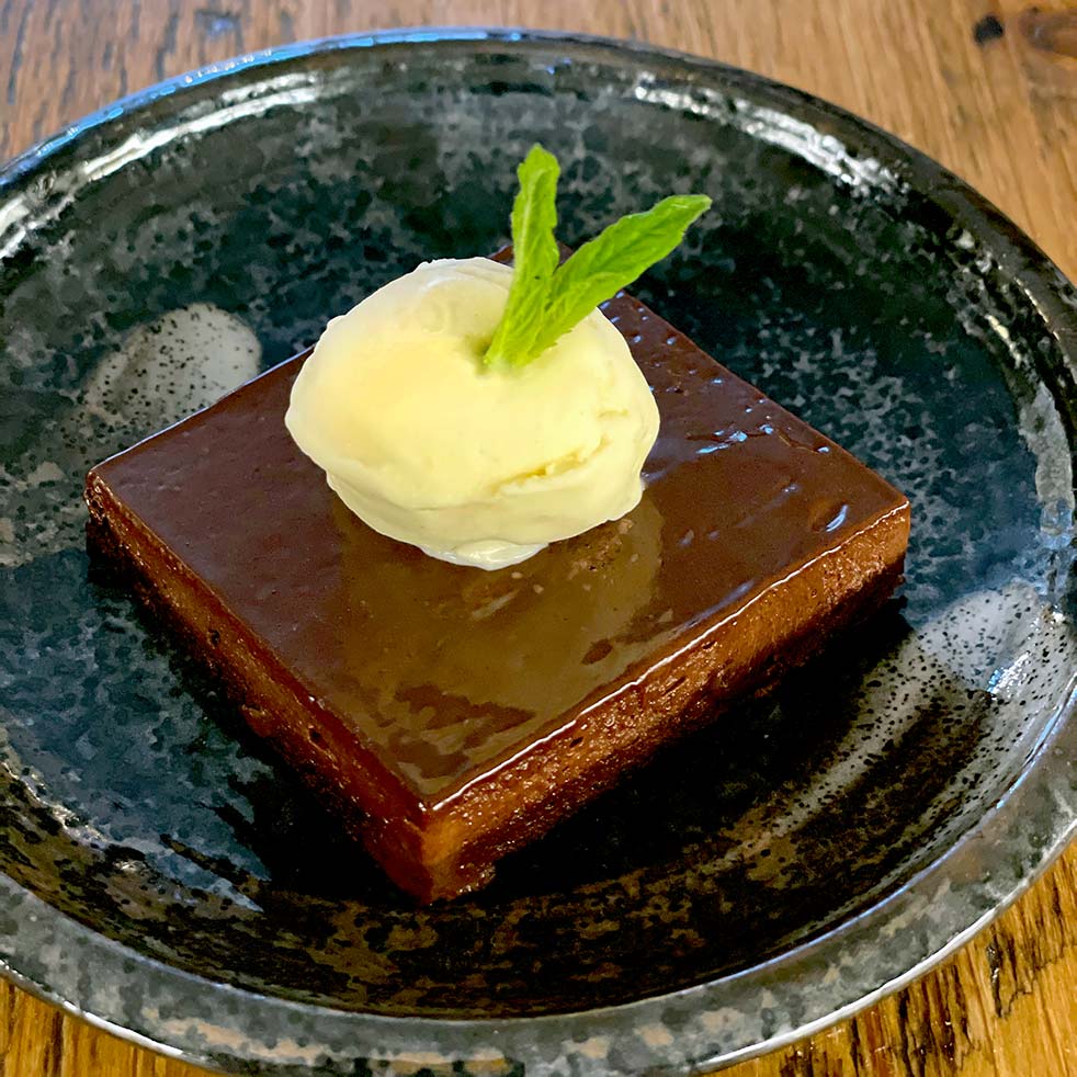 A square slice of chocolate cake topped with a scoop of vanilla ice cream and a fresh mint leaf, served on a dark ceramic plate with a rustic wooden background.
