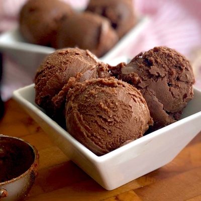 Three scoops of chocolate ice cream in a white square bowl, with more scoops and an ice cream scoop in the background on a wooden surface.