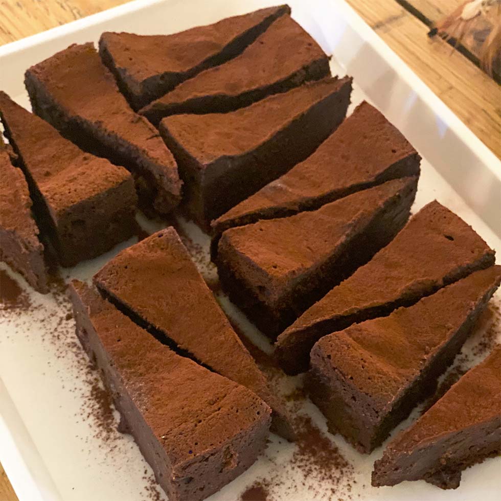 Slices of rich, chocolate cake dusted with cocoa powder are arranged neatly on a white rectangular plate on a wooden table.