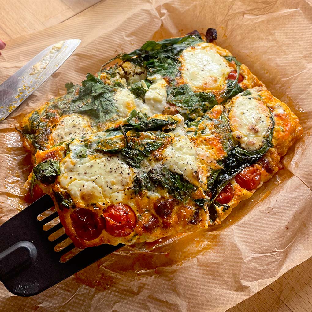 A rectangular baked frittata with spinach, cherry tomatoes, and melted cheese sits on baking parchment. A spatula is lifting a slice, and a knife rests beside it. The dish appears golden and freshly baked.