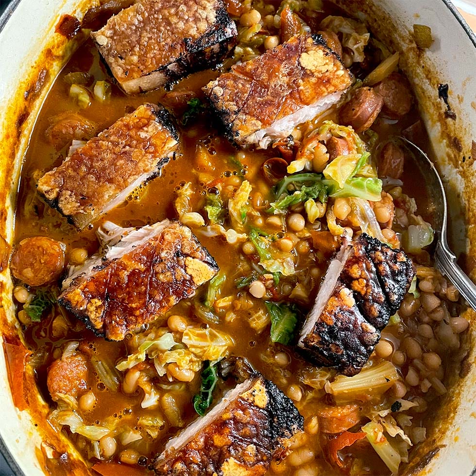 A pot filled with crispy roast pork belly pieces on top of a hearty stew with beans, sausage slices, chopped cabbage, and a rich, tomato-based broth. A spoon rests in the stew.