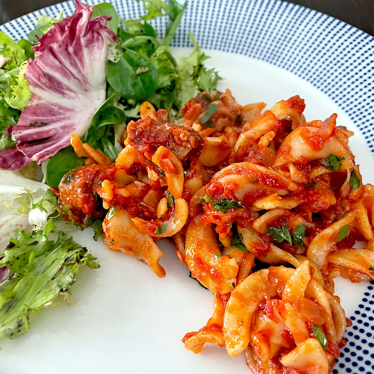 A plate with a portion of marinated seafood salad in red sauce, garnished with herbs, served alongside a mixed green salad with leafy greens.