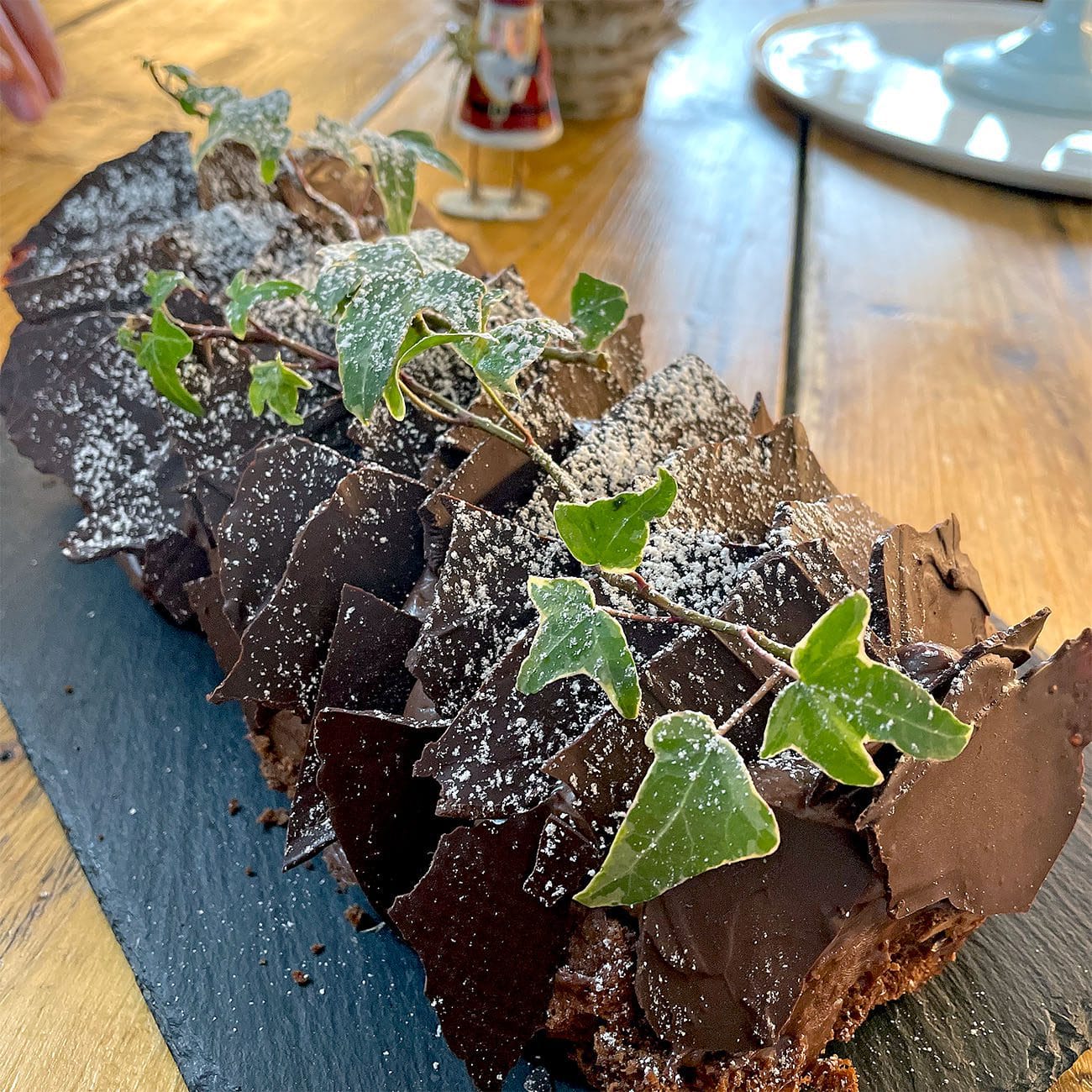 A chocolate yule log cake decorated with ivy leaves and dusted with icing sugar sits on a black slate platter atop a wooden table.