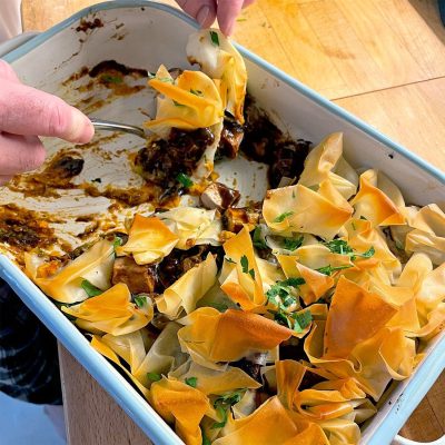 Hands serving a portion of a baked dish with crispy, golden filo pastry on top, garnished with chopped parsley, from a rectangular baking dish on a wooden table. Some filling and pastry remain in the dish.