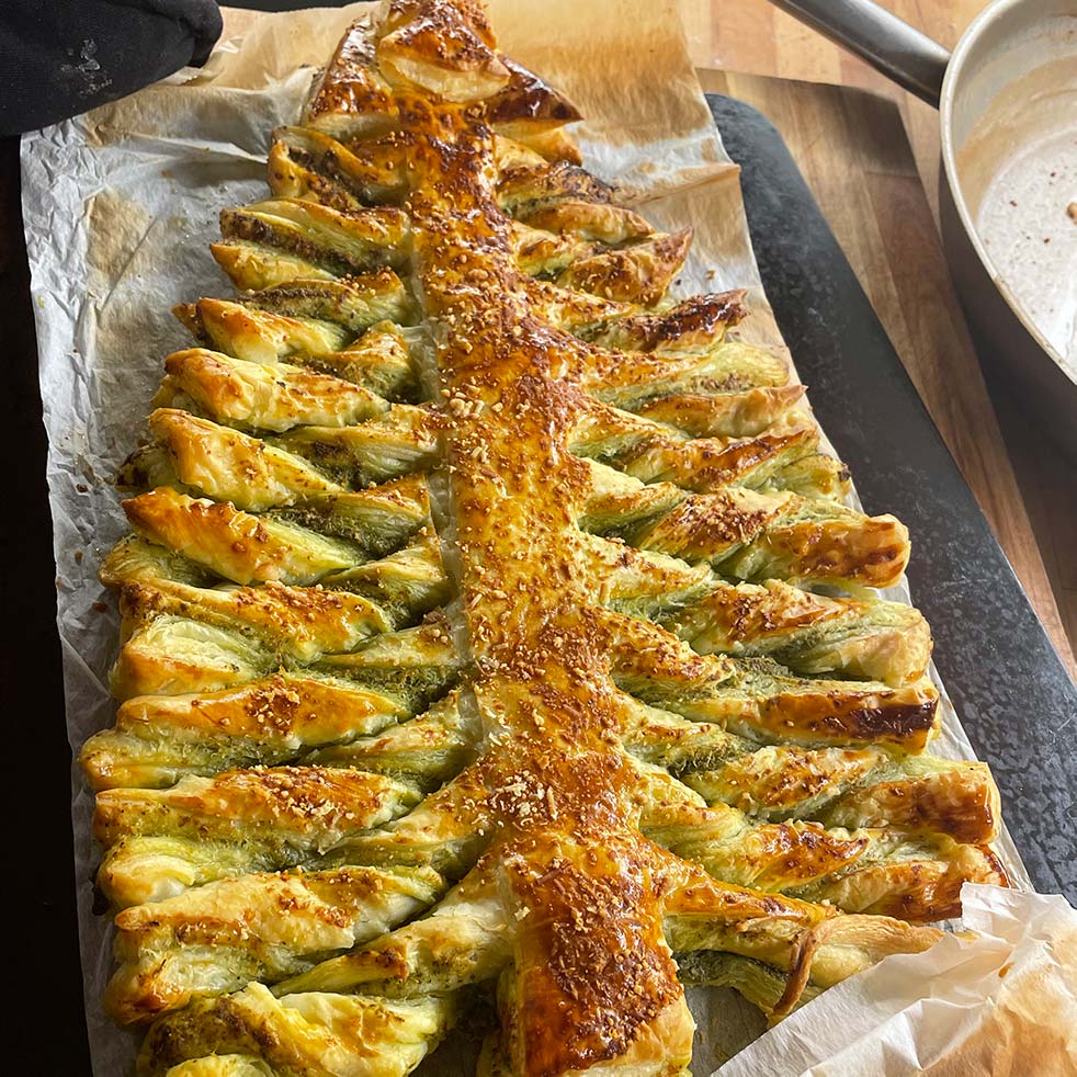 A baked puff pastry shaped like a Christmas tree, with twisted branches and sprinkled with sesame seeds, sits on baking parchment atop a wooden surface.