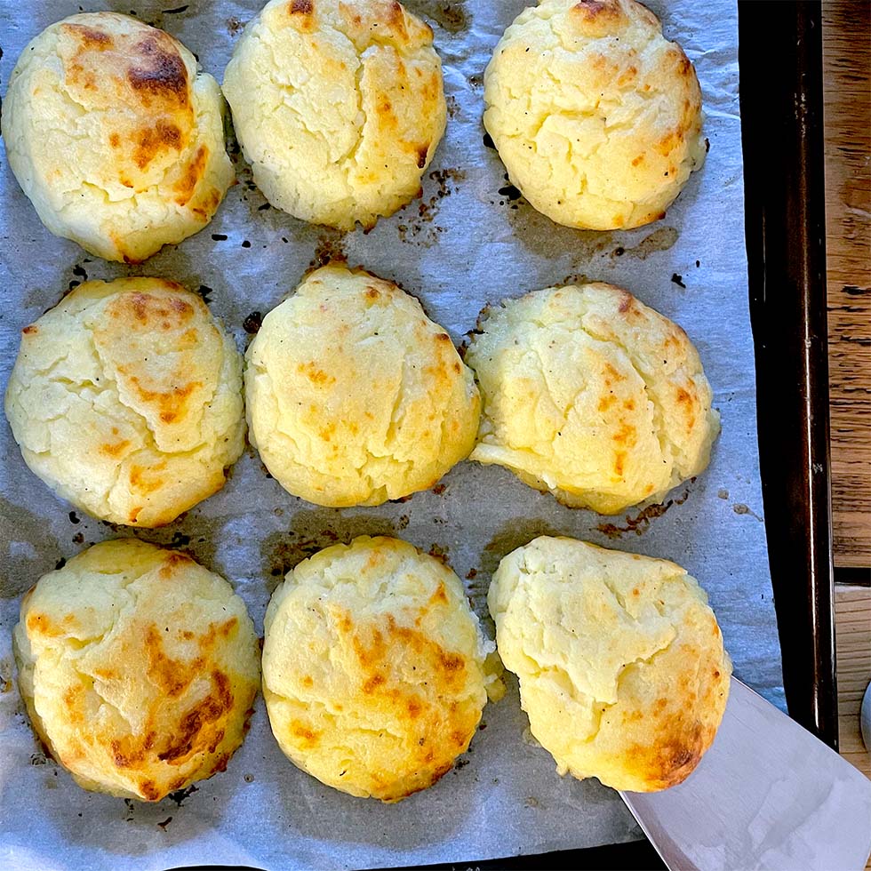 Nine golden-brown, fluffy baked scones are arranged on baking parchment in a baking tray. One scone is being lifted with a spatula, showing its light, airy texture.