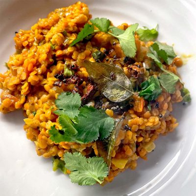 A serving of orange lentil dahl garnished with fresh coriander leaves and bay leaves, plated on a white dish. The dahl appears creamy with visible spices and herbs mixed in.