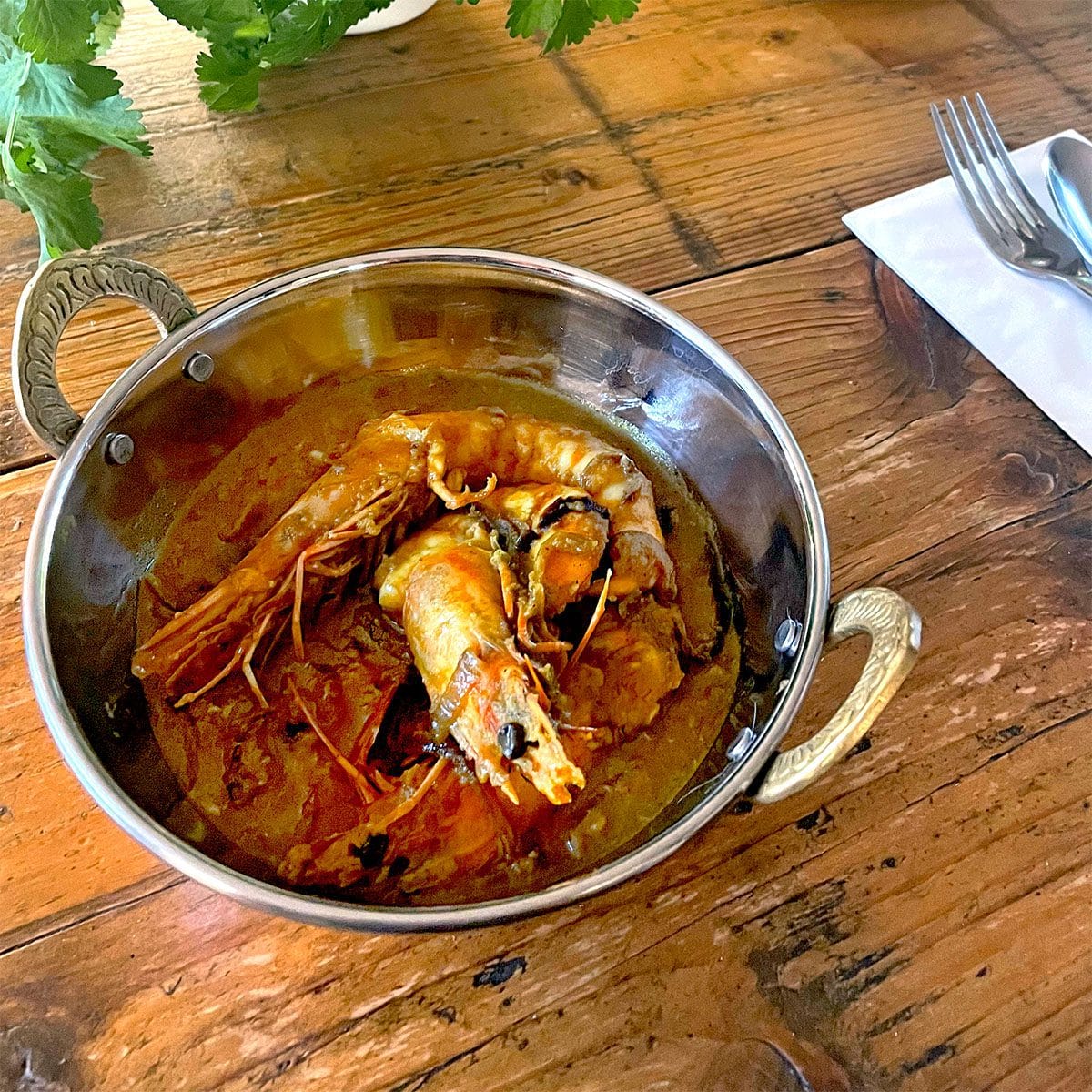 A metal bowl with ornate handles holds a prawn curry in a rich, reddish sauce. The bowl sits on a rustic wooden table beside a folded serviette with a fork and spoon, and some fresh herbs are visible nearby.