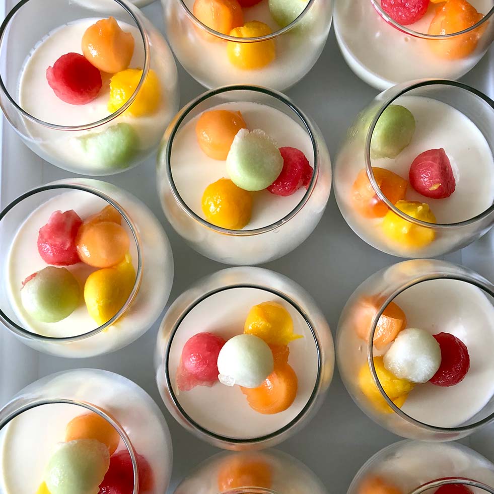 Several glasses filled with a creamy white dessert are topped with colourful melon balls in shades of orange, yellow, red, and green, arranged neatly on a white surface.