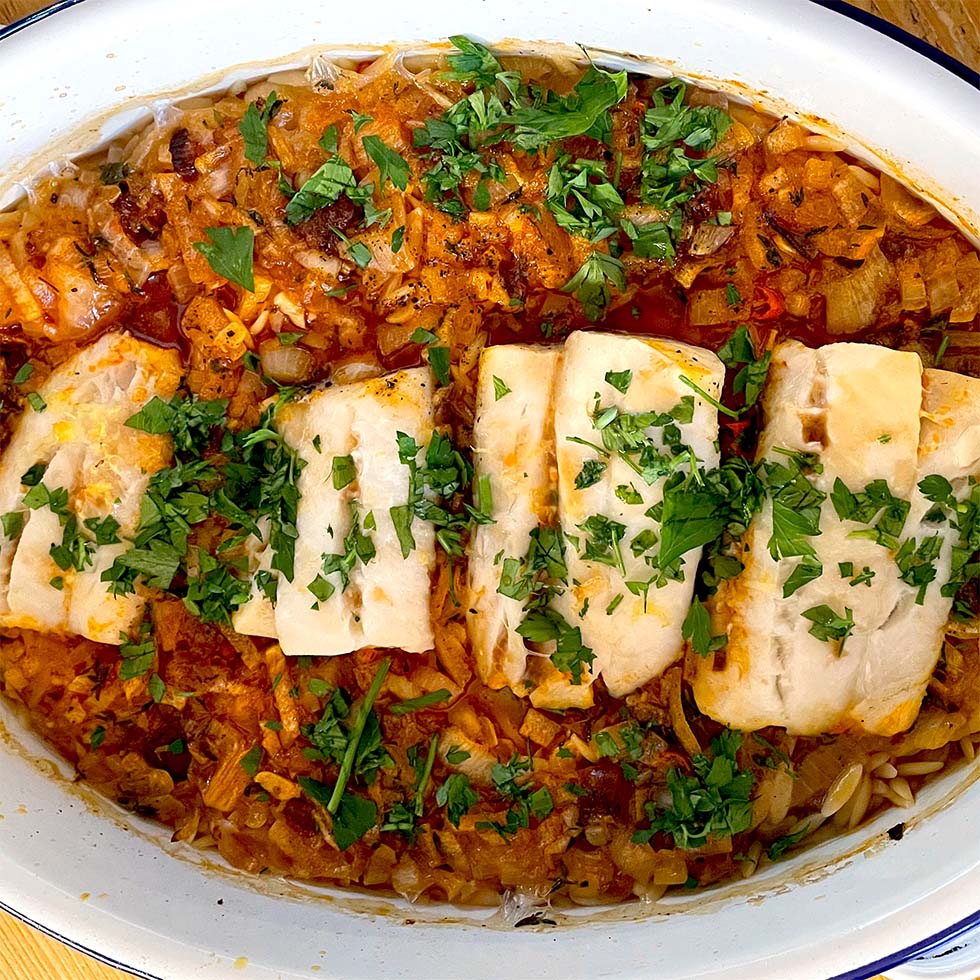 A baked dish featuring four pieces of white fish fillet on a bed of seasoned orzo pasta, topped with chopped fresh herbs, all served in a white oval baking dish.