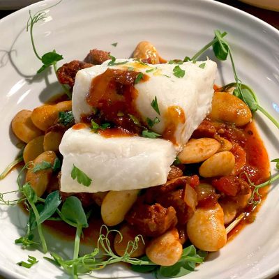 A dish featuring a piece of white fish atop a stew of large white beans, tomato-based sauce, and herbs, garnished with fresh greens on a white plate.
