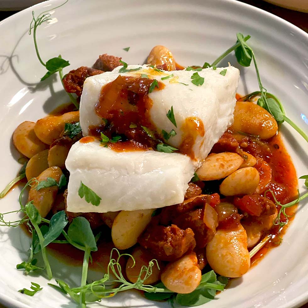 A dish featuring a piece of white fish atop a stew of large white beans, tomato-based sauce, and herbs, garnished with fresh greens on a white plate.