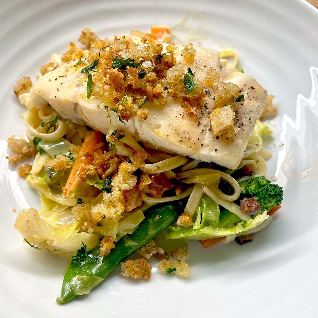 A piece of white fish topped with crispy breadcrumbs sits on a bed of mixed vegetables and pasta, served on a white plate.