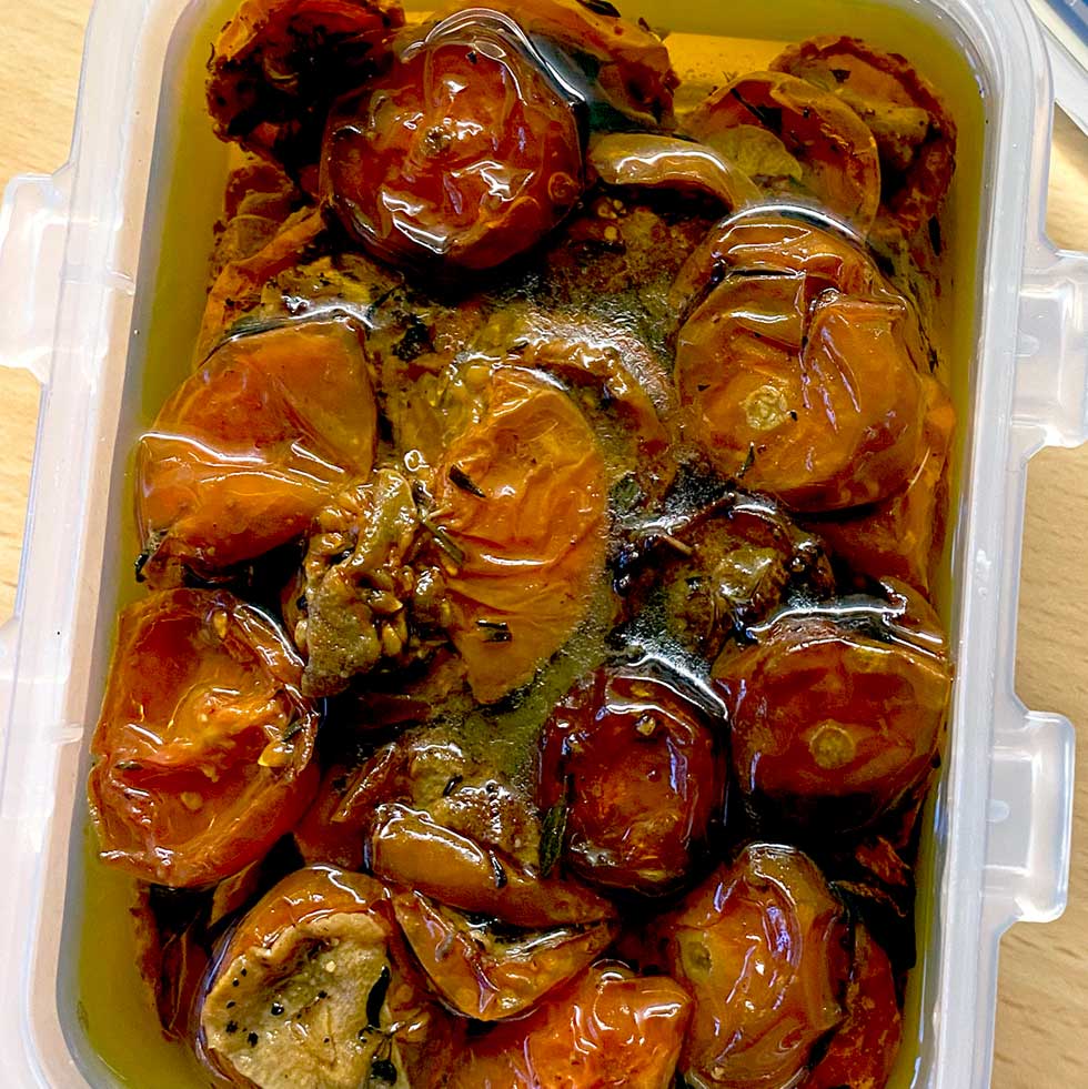 A plastic container filled with slow-roasted tomatoes covered in olive oil, showing shrivelled, glossy red tomato halves and seasoning.